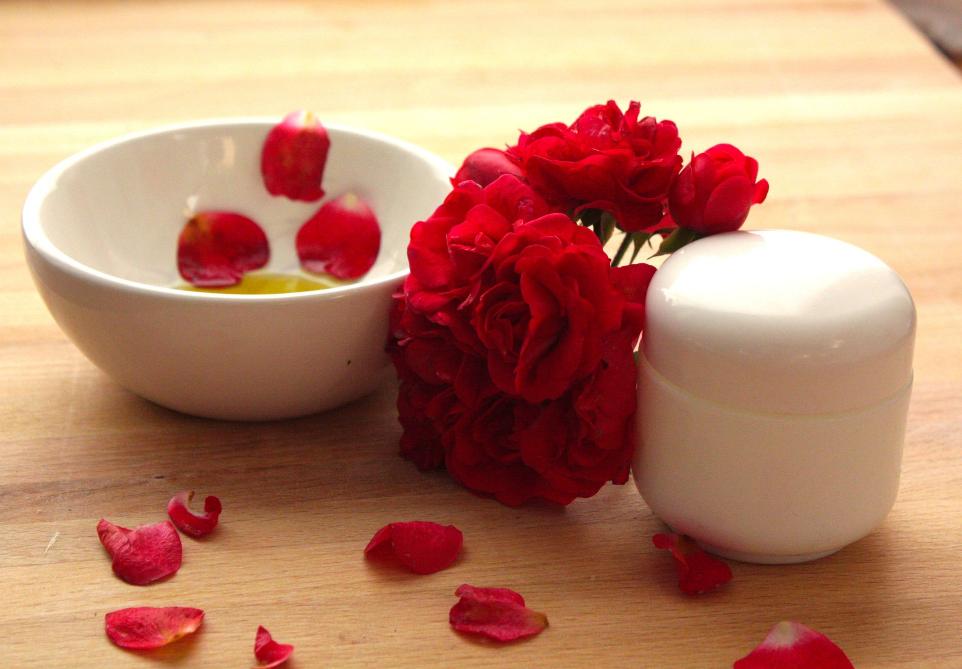 rosenöl in einem weißen keramikschaelchen mit rosenblütenblättern und roten rosen, davor ein weißer cremetigel