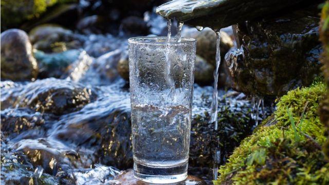 kraeuterblume-blog-darmgesundheit-trinken Glas auf Steinen einer natürlichen Quelle, frisches Quellwasser tropft ins Glas